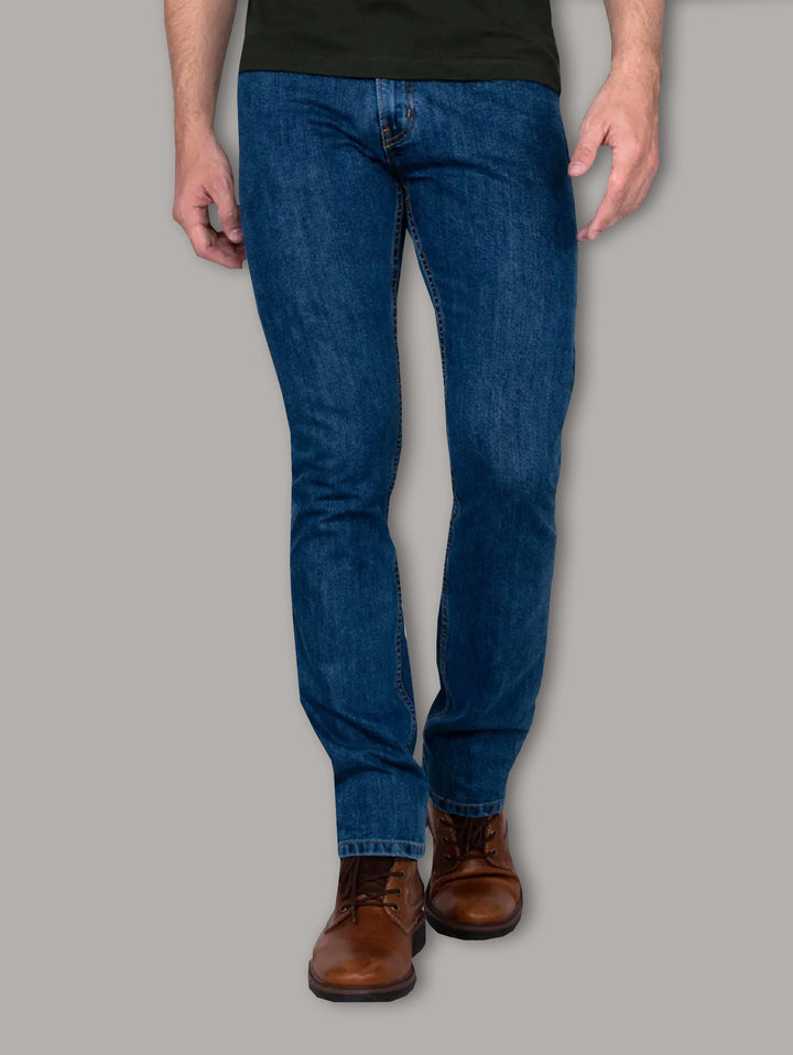 Hombre vistiendo jeans azul oscuro OGGI y botas color café sobre fondo gris.