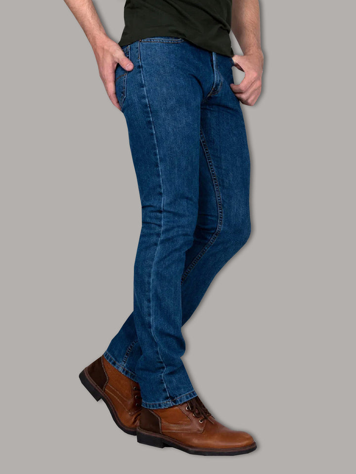 Hombre vistiendo jeans azul oscuro OGGI y botas color café sobre fondo gris.