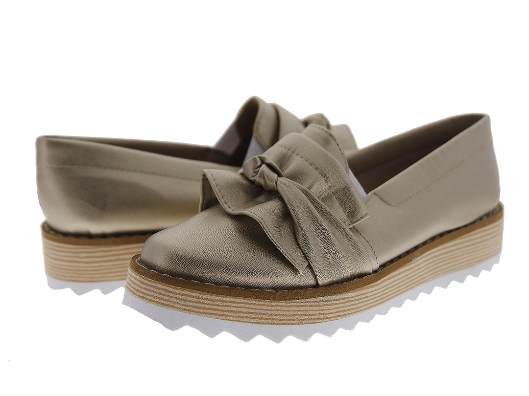 Zapatos mocasín color dorado metálico con moño frontal y suela de plataforma blanca y café.