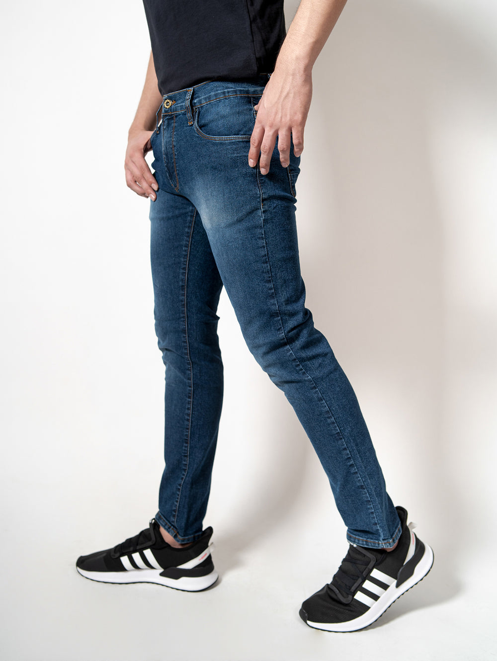 Hombre vistiendo pantalones de mezclilla azules ajustados y calzado deportivo negro sobre fondo blanco.