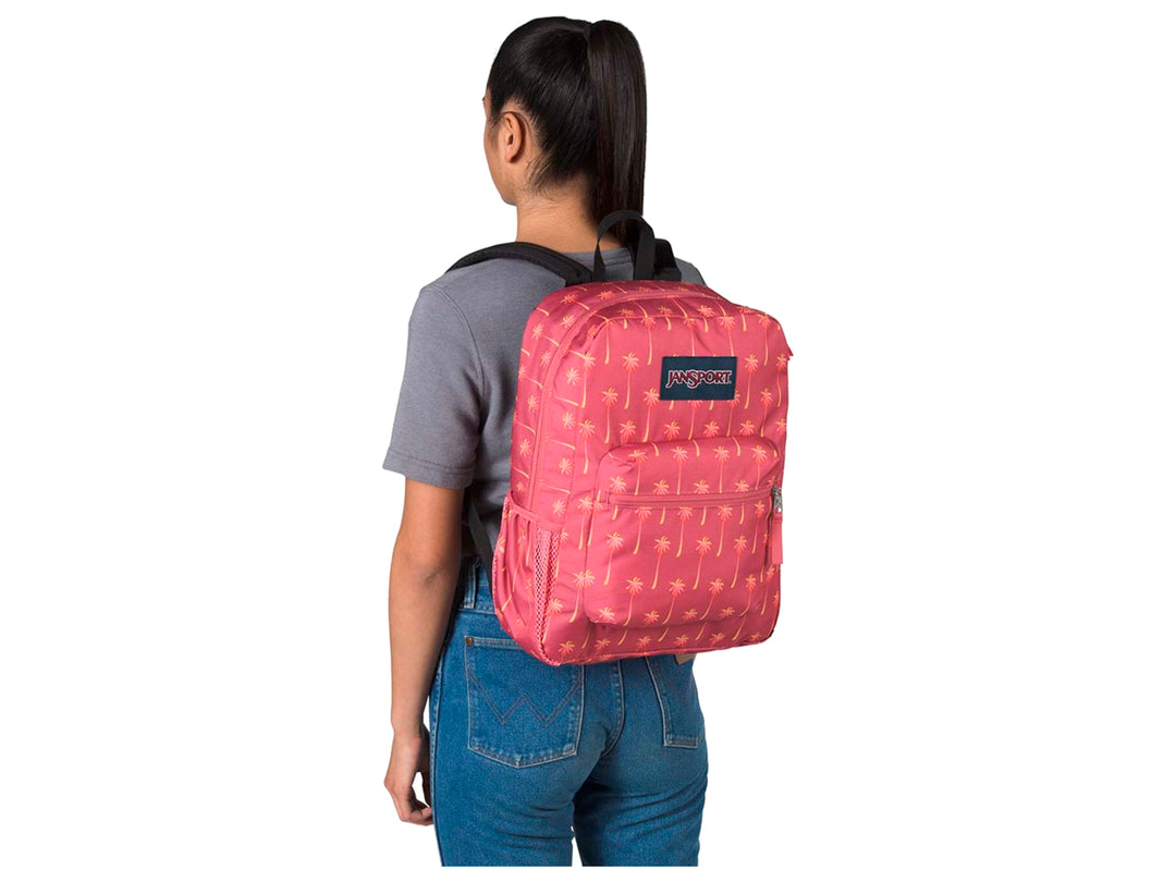Mochila JanSport rosa con estampado de palmeras amarillas y un termo morado en el bolsillo lateral.