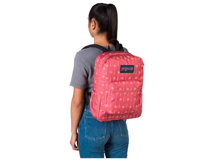 Mochila JanSport rosa con estampado de palmeras amarillas y un termo morado en el bolsillo lateral.