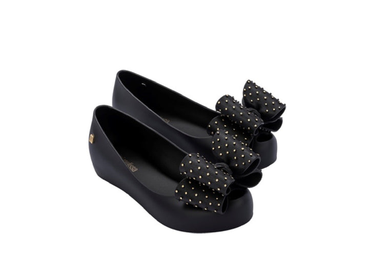 Zapatos planos negros para niña con moño decorado con puntos dorados.
