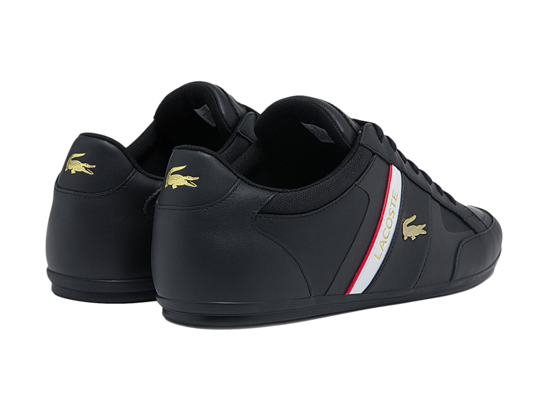Tenis Lacoste color negro con franja blanca y roja lateral y logotipo del cocodrilo.