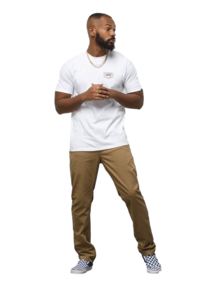Hombre vistiendo pantalón chino color café marca Vans con camiseta blanca y tenis a cuadros.
