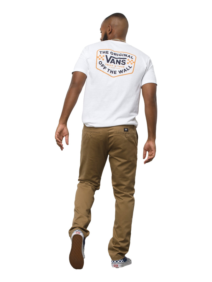 Hombre vistiendo pantalón chino color café marca Vans con camiseta blanca y tenis a cuadros.