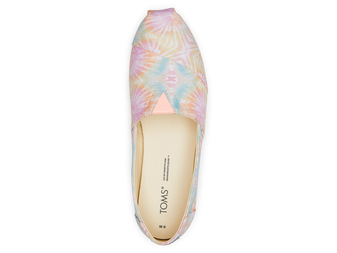 Zapato TOMS estilo slip-on de tela con estampado tie dye en tonos rosa, amarillo y azul con suela blanca.