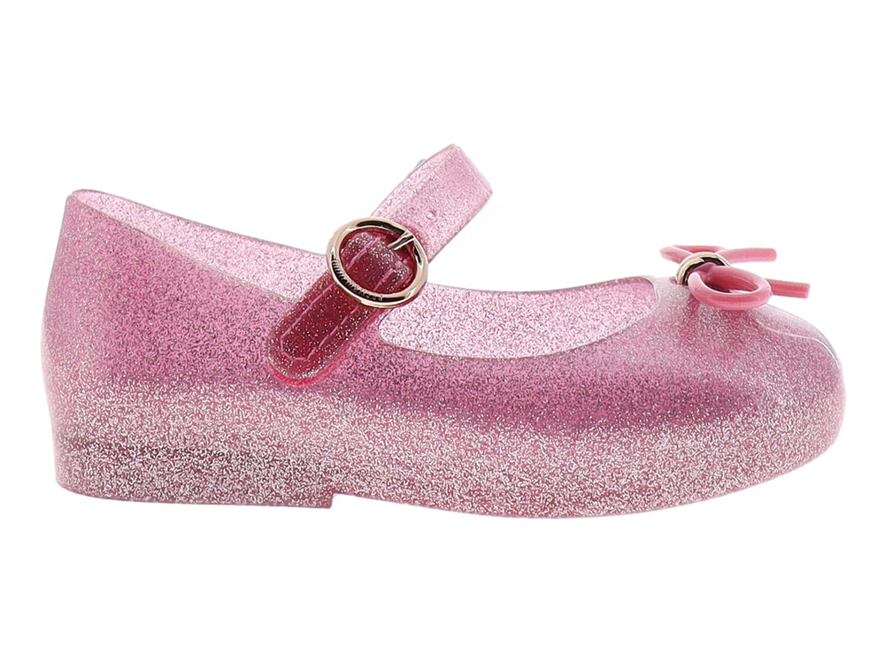Zapato infantil Mini Melissa color rosa con glitter, moño decorativo y correa con hebilla dorada.