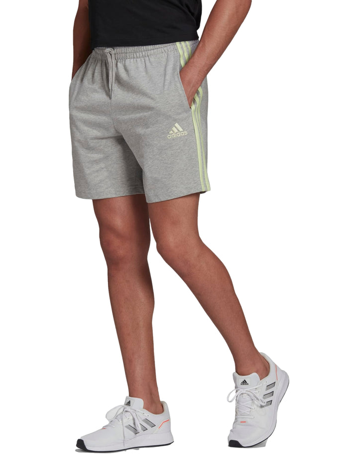 Hombre vistiendo pantalones cortos deportivos Adidas color gris con rayas laterales verde lima.