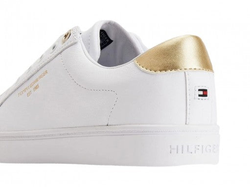Tenis blancos Tommy Hilfiger estilo slip-on con detalle dorado en el talón y suela blanca.
