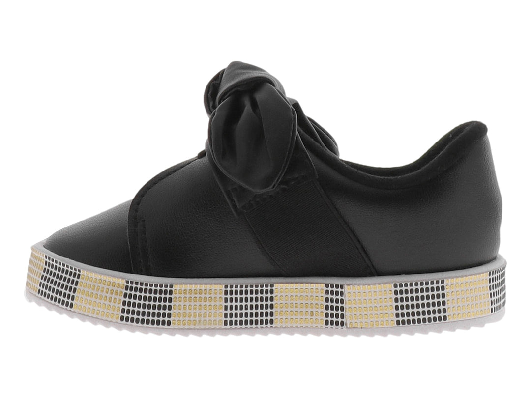 Zapato negro para niña con moño decorativo y suela con patrón de cuadros negros y dorados.