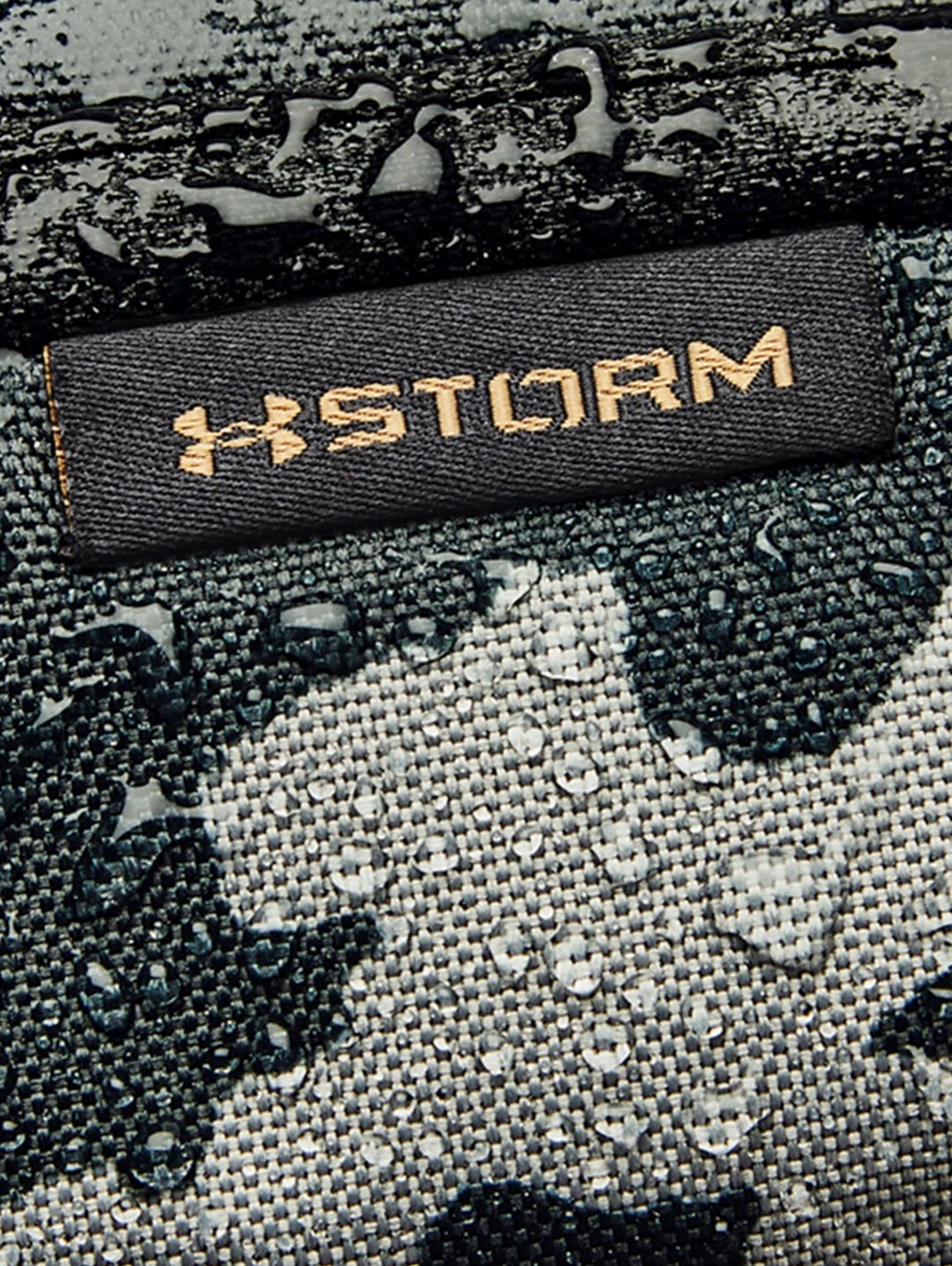 Mochila Under Armour Hustle 5.0 con estampado de camuflaje en tonos negro y gris con logos dorados.