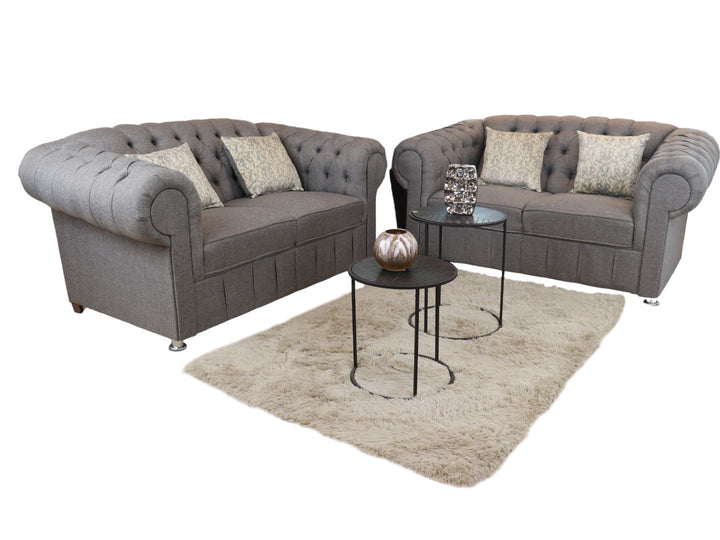 Juego de sala compuesto por dos sofás grises estilo Chesterfield con capitoné y cojines decorativos.