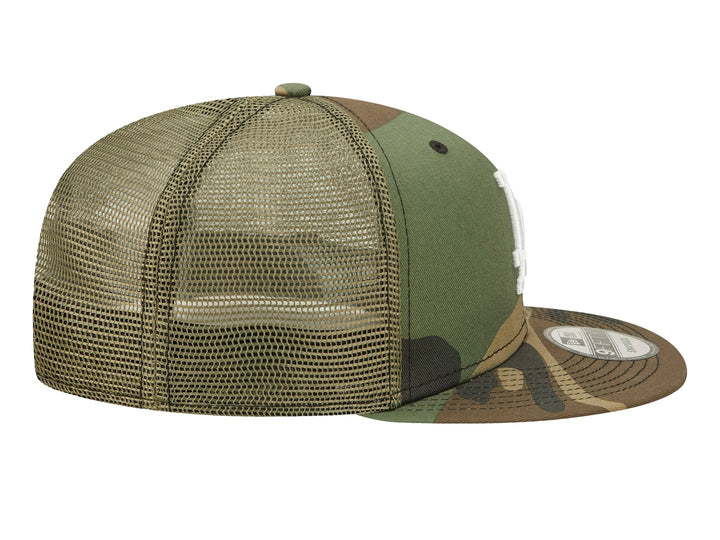 Gorra New Era con estampado de camuflaje en tonos verdes y cafés, visera plana y logo LA bordado en blanco.