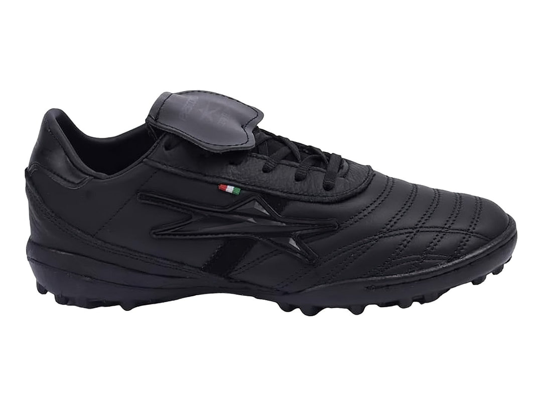 Zapato de fútbol negro con lengüeta plegable y suela para césped sintético.