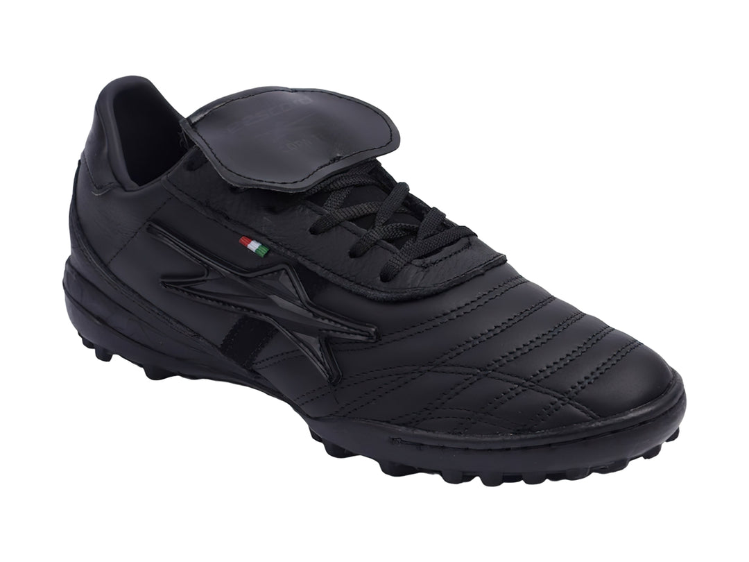 Zapato de fútbol negro con lengüeta plegable y suela para césped sintético.