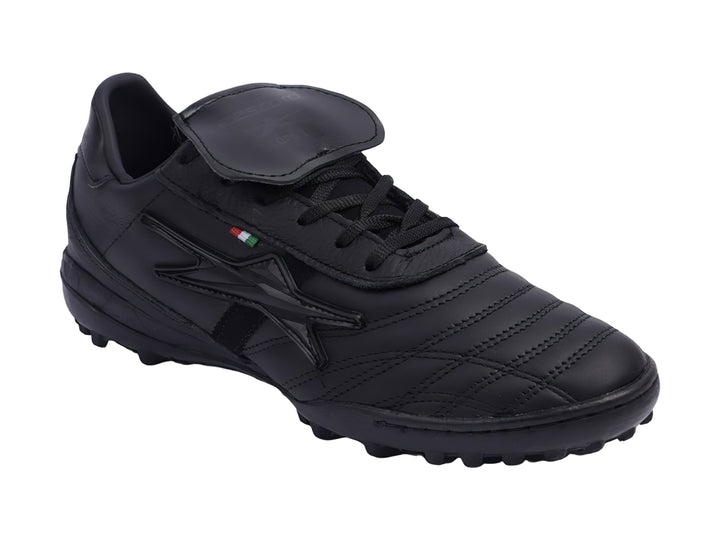 Zapato de fútbol negro con lengüeta plegable y suela para césped sintético.
