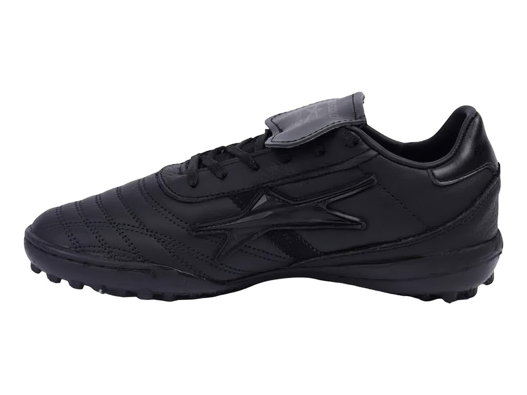 Zapato de fútbol negro con lengüeta plegable y suela para césped sintético.