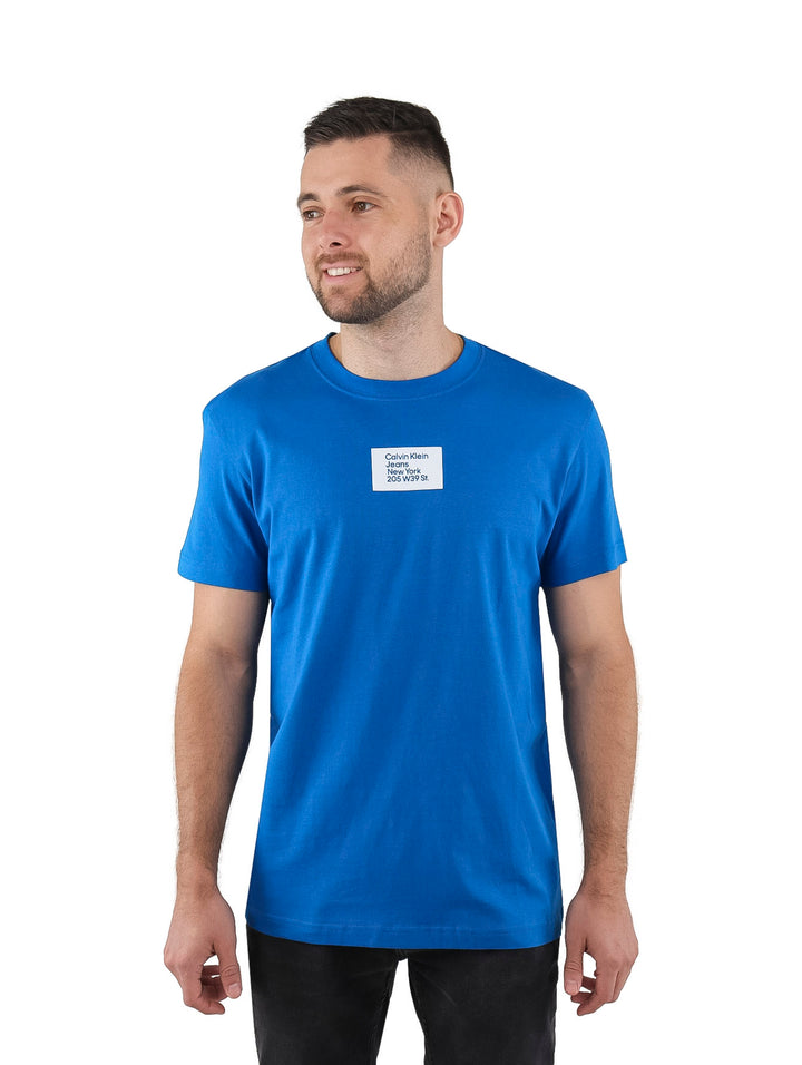 Hombre vistiendo una camiseta azul de manga corta con un pequeño detalle rectangular blanco en el pecho.