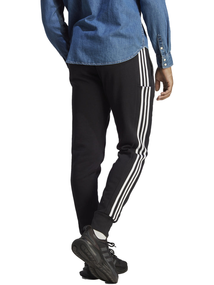 Hombre vistiendo pantalones deportivos Adidas color negro con tres franjas blancas laterales y logo en la pierna.