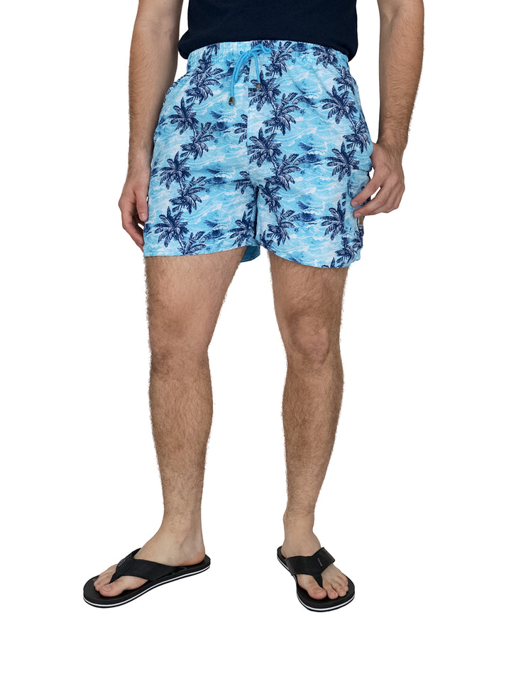 Hombre usando un traje de baño azul con estampado de palmeras y sandalias negras.