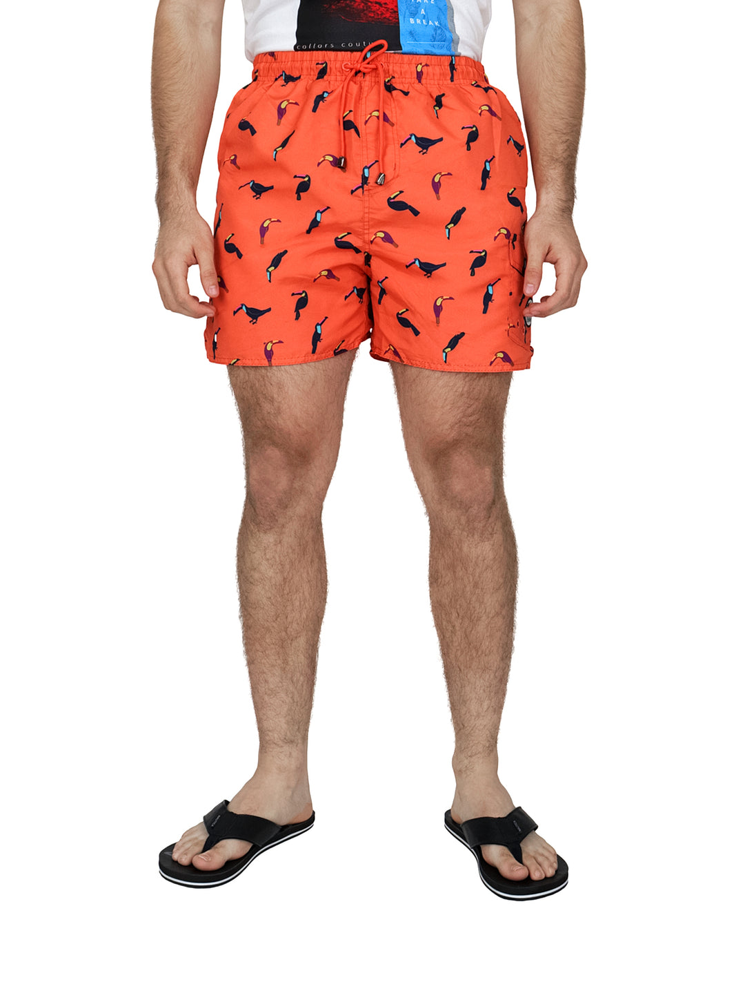 Hombre vistiendo un traje de baño corto color coral con estampado de tucanes negros y sandalias negras.