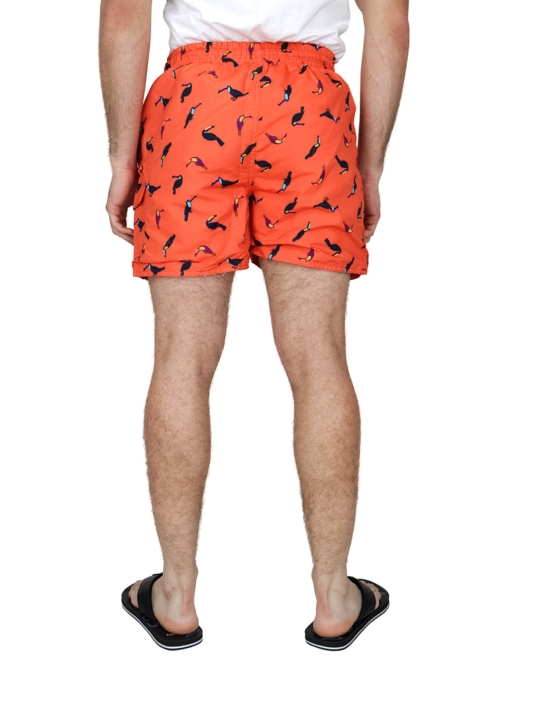 Hombre vistiendo un traje de baño corto color coral con estampado de tucanes negros y sandalias negras.
