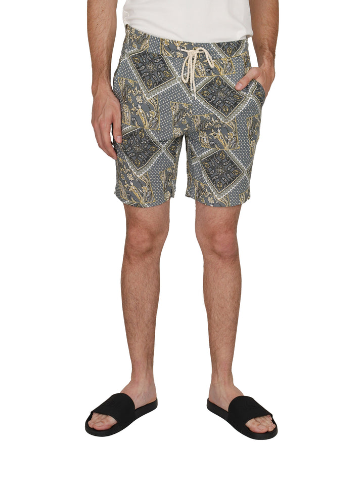 Hombre vistiendo shorts grises con estampado étnico amarillo y blanco, camiseta blanca y sandalias negras.