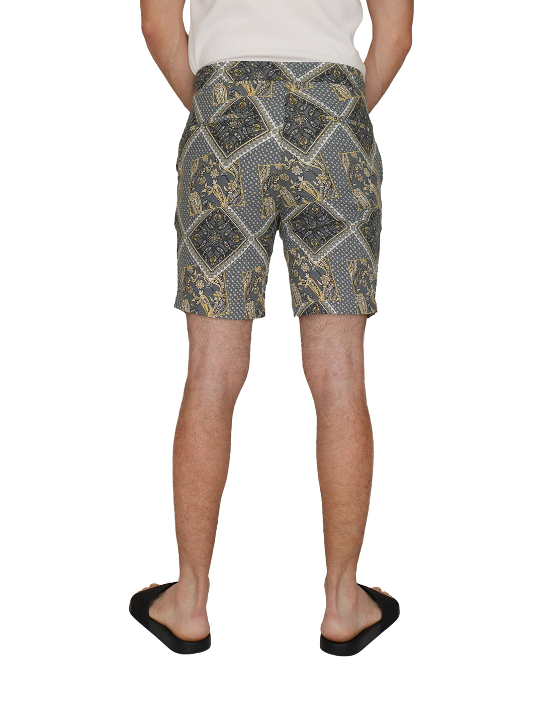 Hombre vistiendo shorts grises con estampado étnico amarillo y blanco, camiseta blanca y sandalias negras.