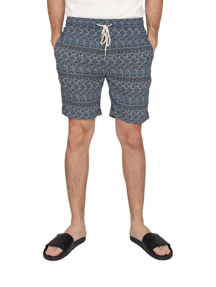 Hombre usando shorts de baño azules con estampado étnico y cordones blancos.
