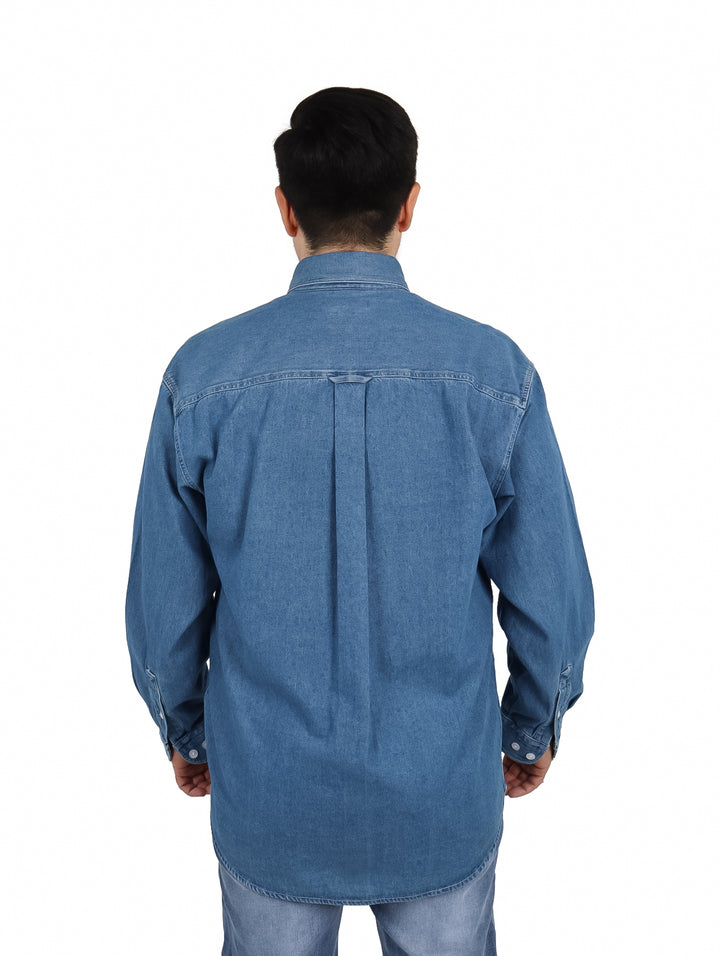 Hombre vistiendo una camisa de mezclilla azul de manga larga con botones blancos y un bolsillo frontal.