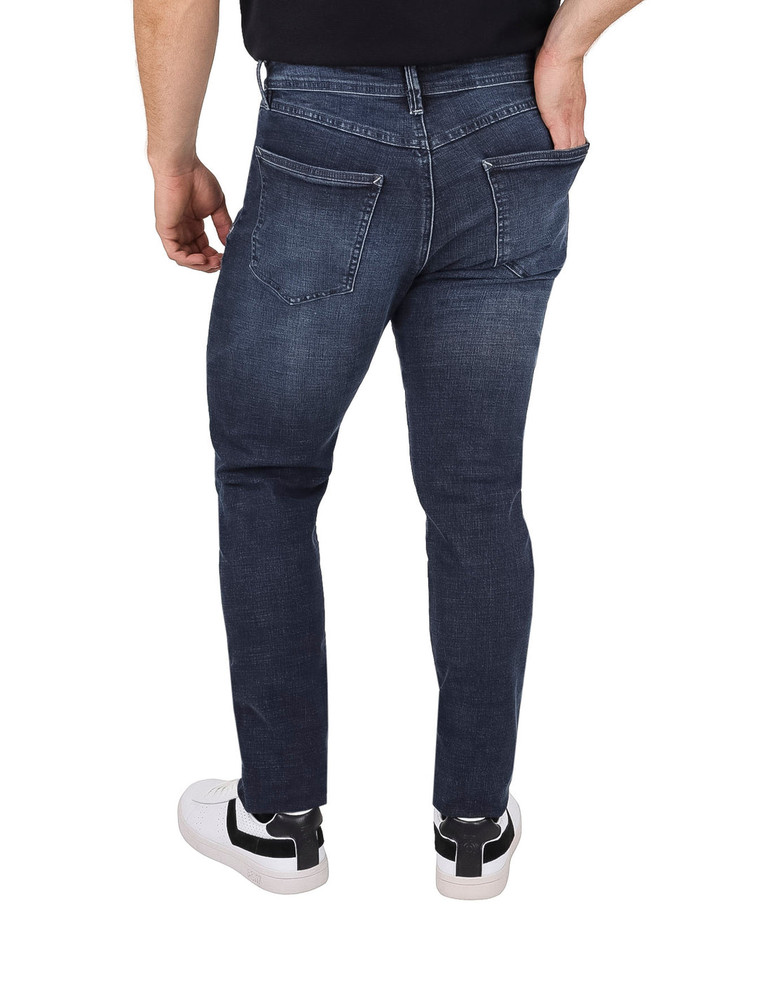 Hombre vistiendo pantalones jeans de color azul oscuro y tenis blancos sobre fondo blanco.