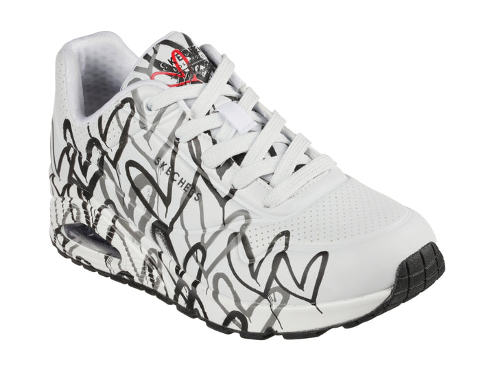 Tenis Skechers blancos con estampado de corazones en negro y gris, diseño deportivo con suela blanca y negra.