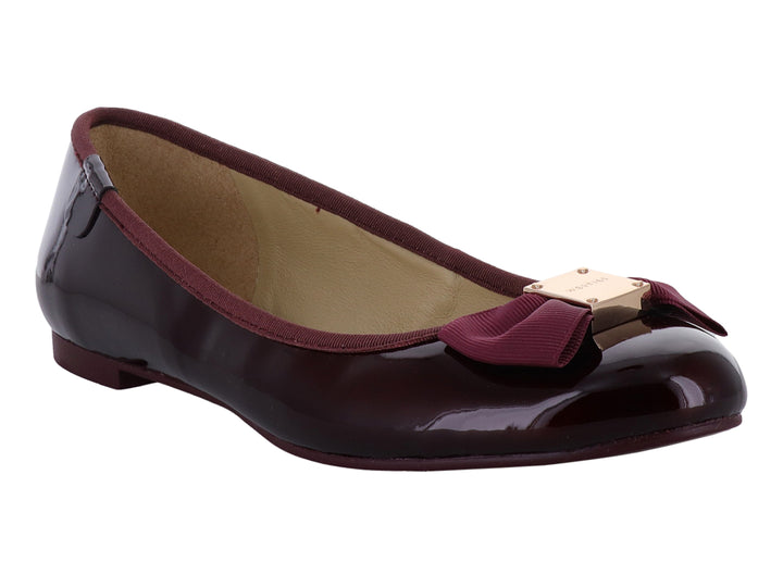 Zapatos planos color wine con acabado brillante, decorados con un moño y herraje dorado en la punta.