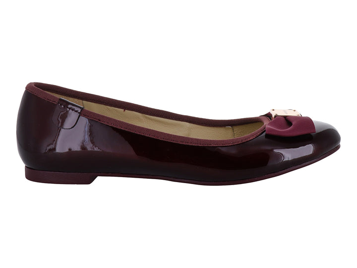 Zapatos planos color wine con acabado brillante, decorados con un moño y herraje dorado en la punta.