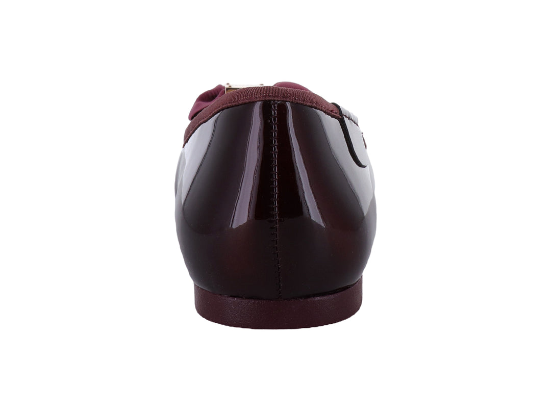 Zapatos planos color wine con acabado brillante, decorados con un moño y herraje dorado en la punta.