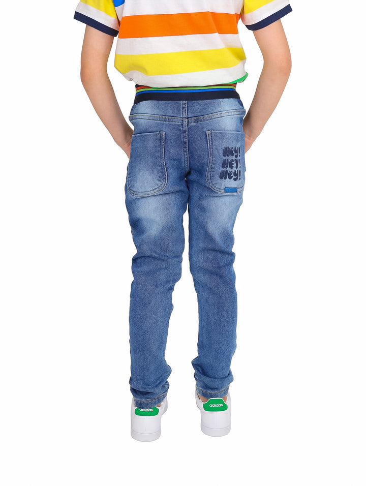 Pantalones vaqueros denim para niño con cintura elástica, un parche decorativo rojo y texto bordado en la pierna.