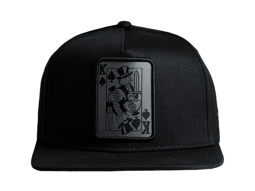 Gorra snapback color negro con un parche frontal rectangular que representa una carta del rey de picas.