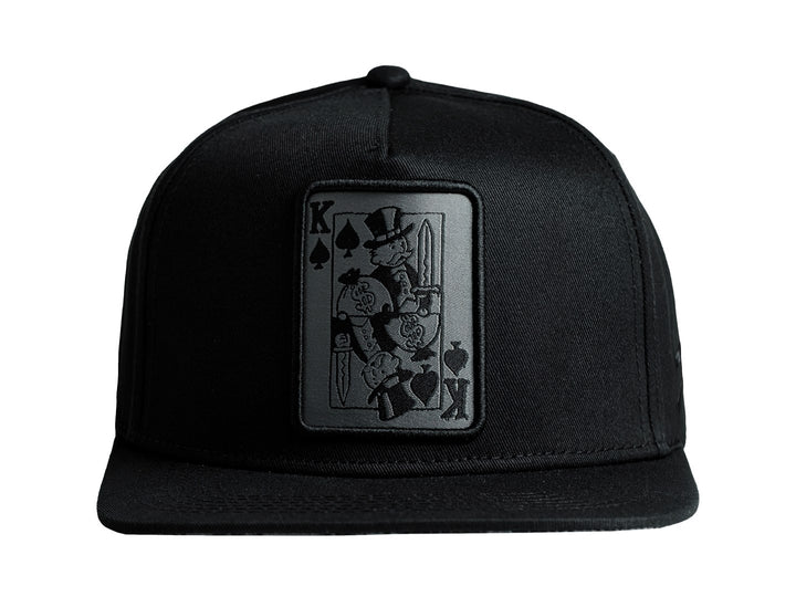 Gorra snapback color negro con un parche frontal rectangular que representa una carta del rey de picas.
