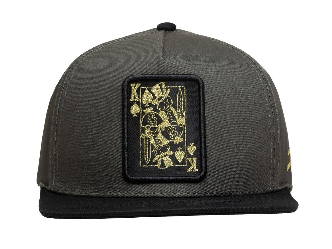 Gorra de béisbol color gris con visera negra y un parche rectangular frontal con diseño de carta del rey en dorado.
