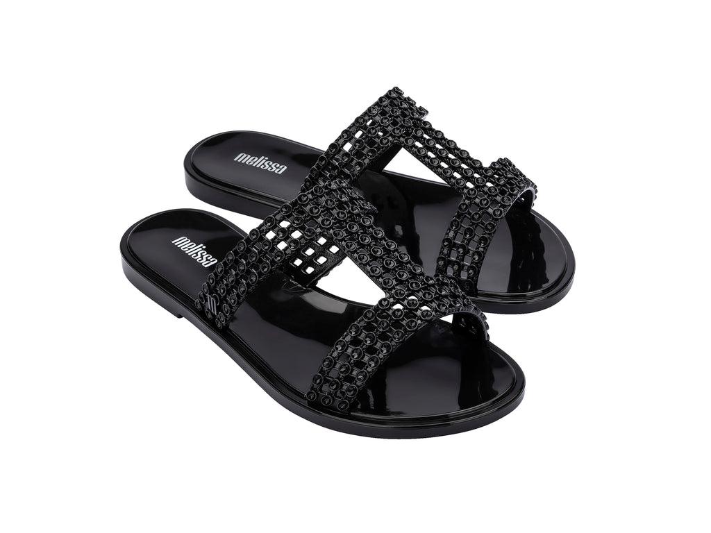 Sandalias Melissa Glowing Slide en color negro brillante con tiras decorativas entrelazadas.