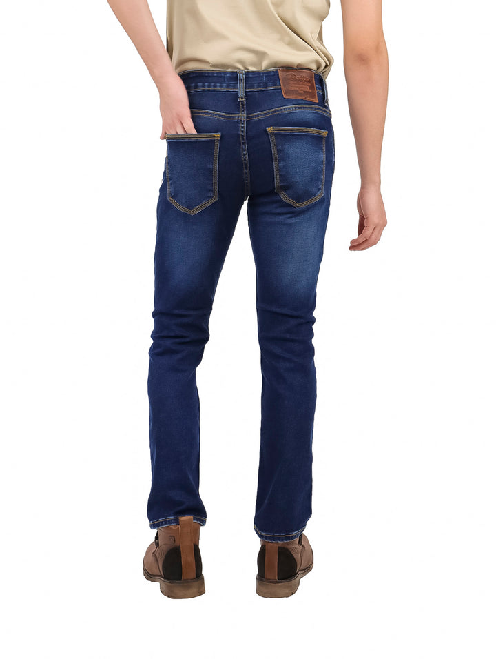 Hombre vistiendo pantalones jeans slim fit color azul oscuro, botas cafés y playera beige.