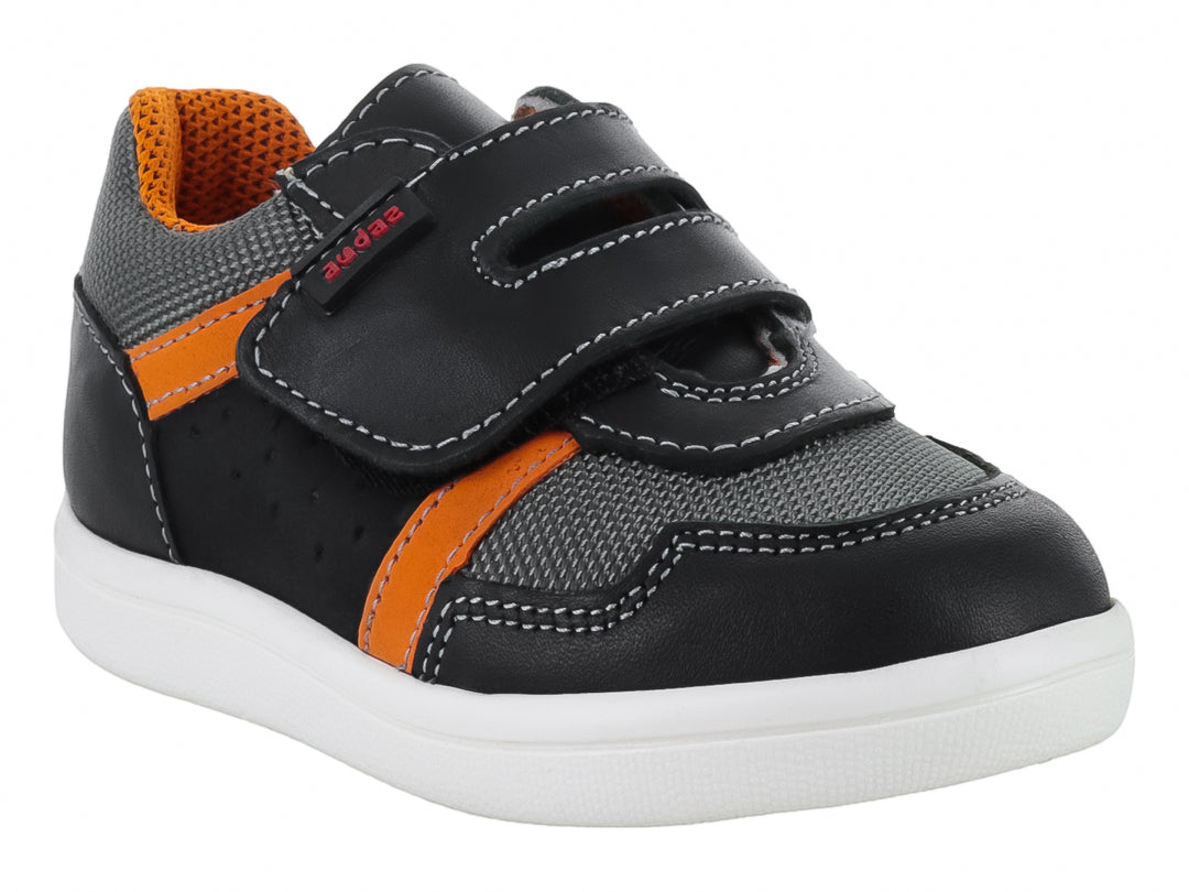 Tenis infantil color negro con detalles en gris y naranja, suela blanca y cierre de velcro.
