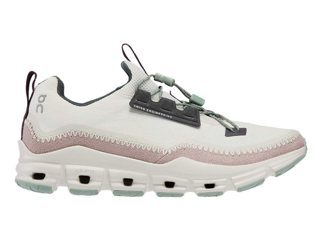 Tenis deportivos On Cloud color gris claro con banda morada y suela blanca con detalles verdes.