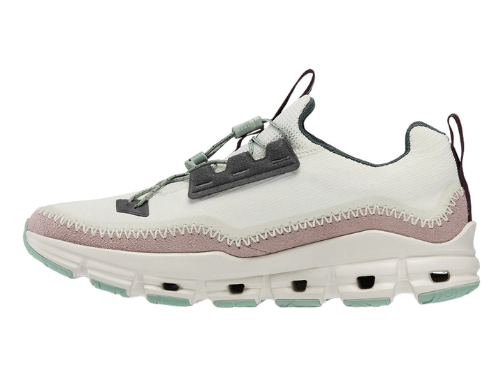 Tenis deportivos On Cloud color gris claro con banda morada y suela blanca con detalles verdes.