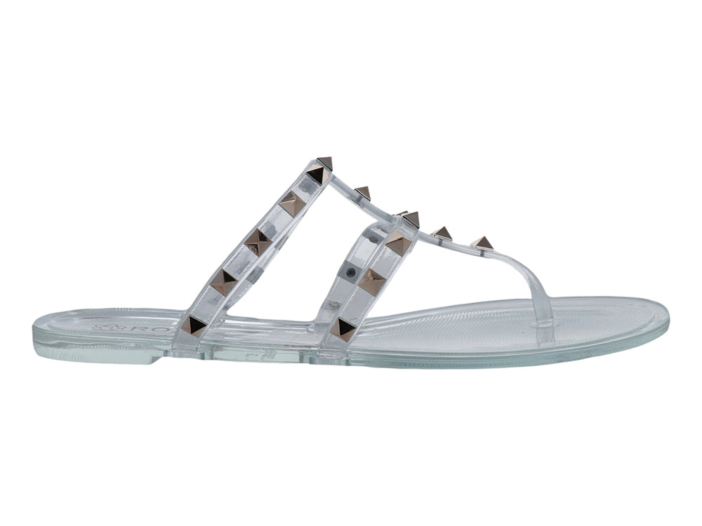 Sandalia plana transparente para niña con tiras decoradas con tachuelas doradas.
