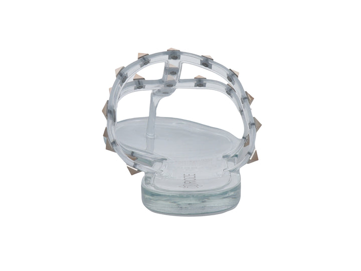 Sandalia plana transparente para niña con tiras decoradas con tachuelas doradas.