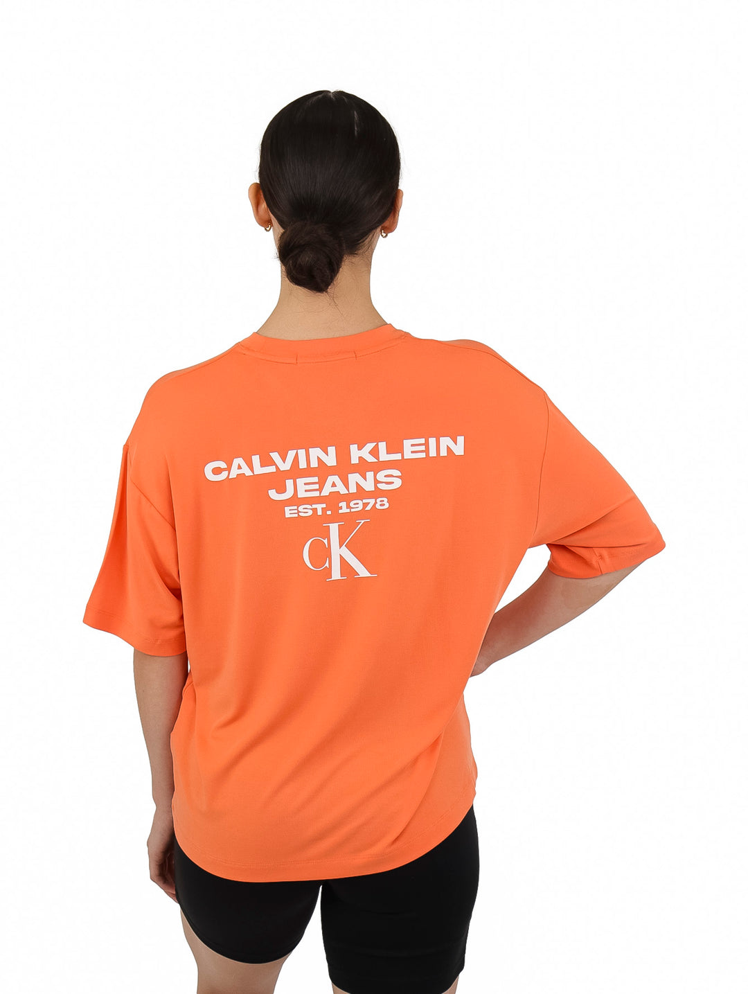 Mujer vistiendo una camiseta naranja holgada de Calvin Klein y shorts negros sobre fondo blanco.