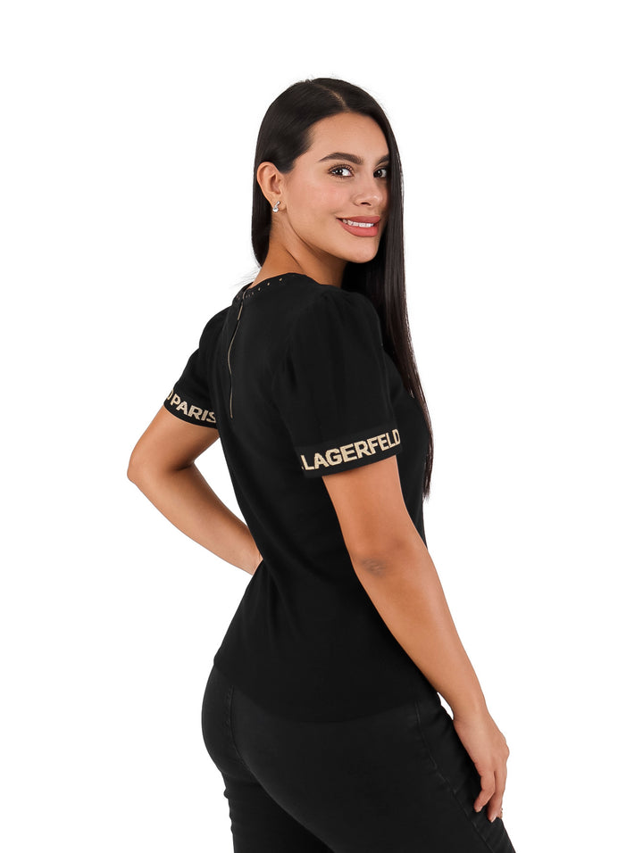 Mujer vistiendo blusa negra de manga corta con letras doradas en las mangas y detalles en el cuello.