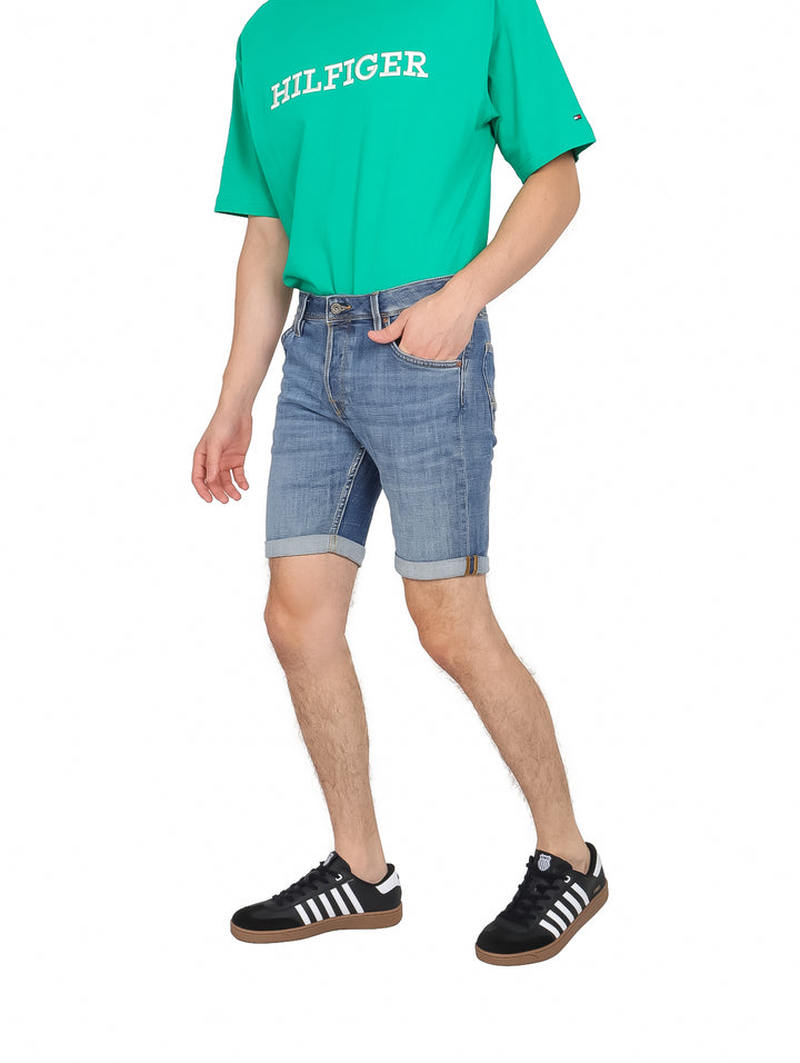 Hombre vistiendo shorts de mezclilla color azul, camiseta verde y tenis negros sobre fondo blanco.